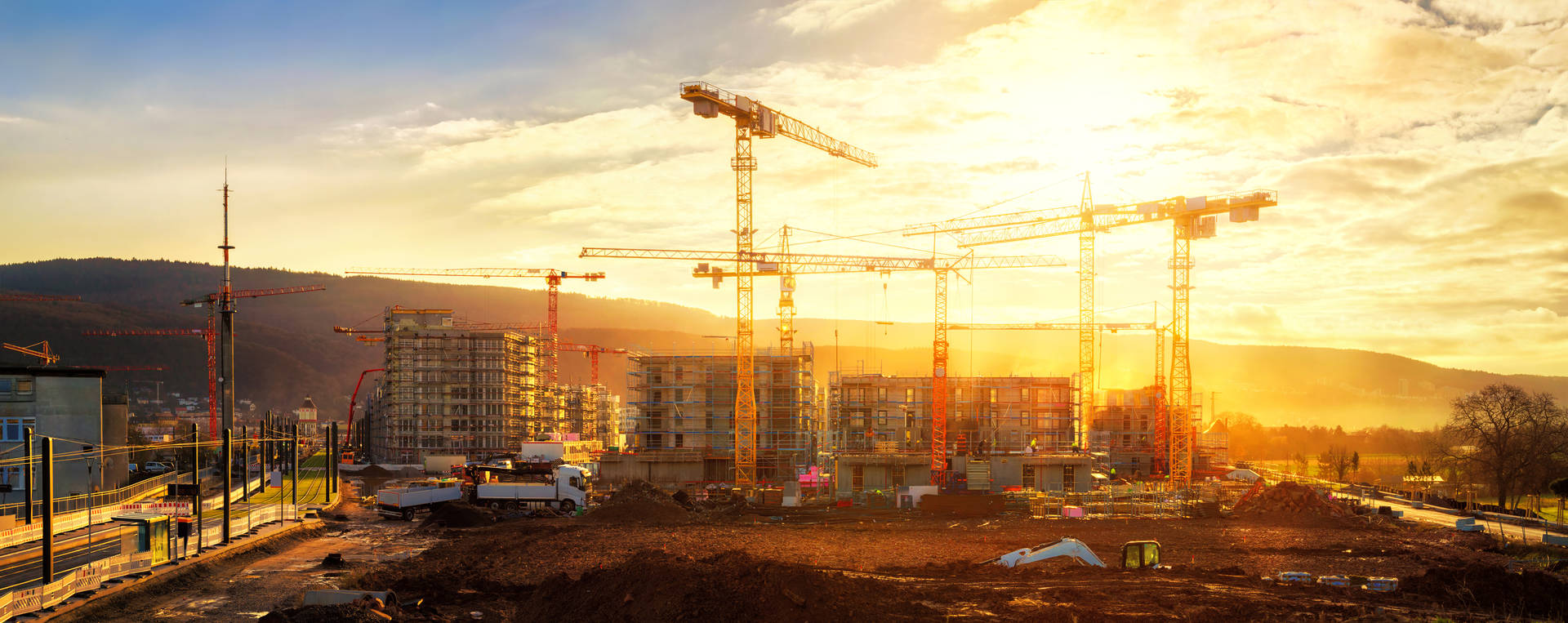 Panoramic view of a construction site at sunset, featuring multiple cranes, partially constructed buildings, and surrounding natural landscapes.
