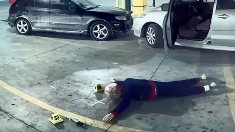A body lying face down on the ground near two vehicles with evidence marked for forensic reconstruction