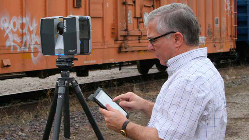 A man using a mobile device and FARO 3D scanning device near a train